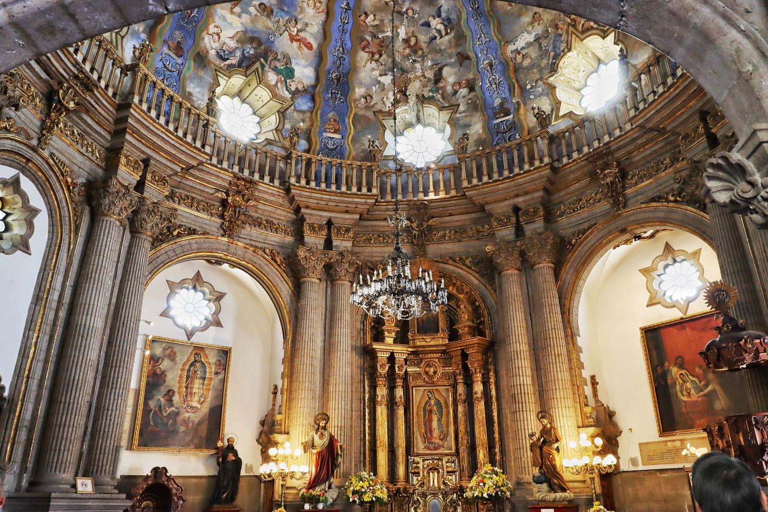Ornate baroque church interior with a painted domed ceiling, hanging chandelier, gilded altarpiece and carved stone columns.