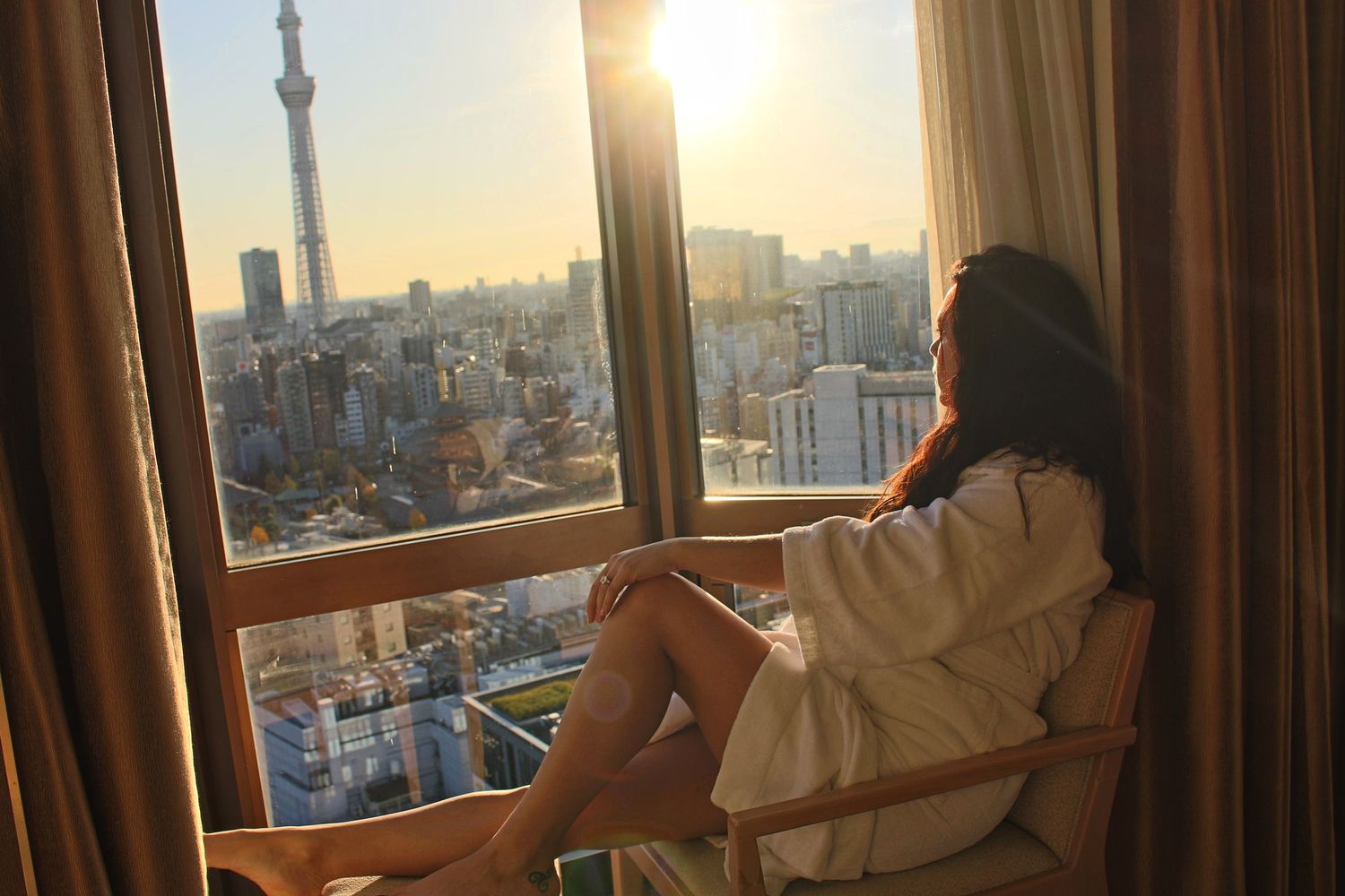 Woman in a white robe relaxes by a hotel window at sunset overlooking the Tokyo skyline and Skytree tower.