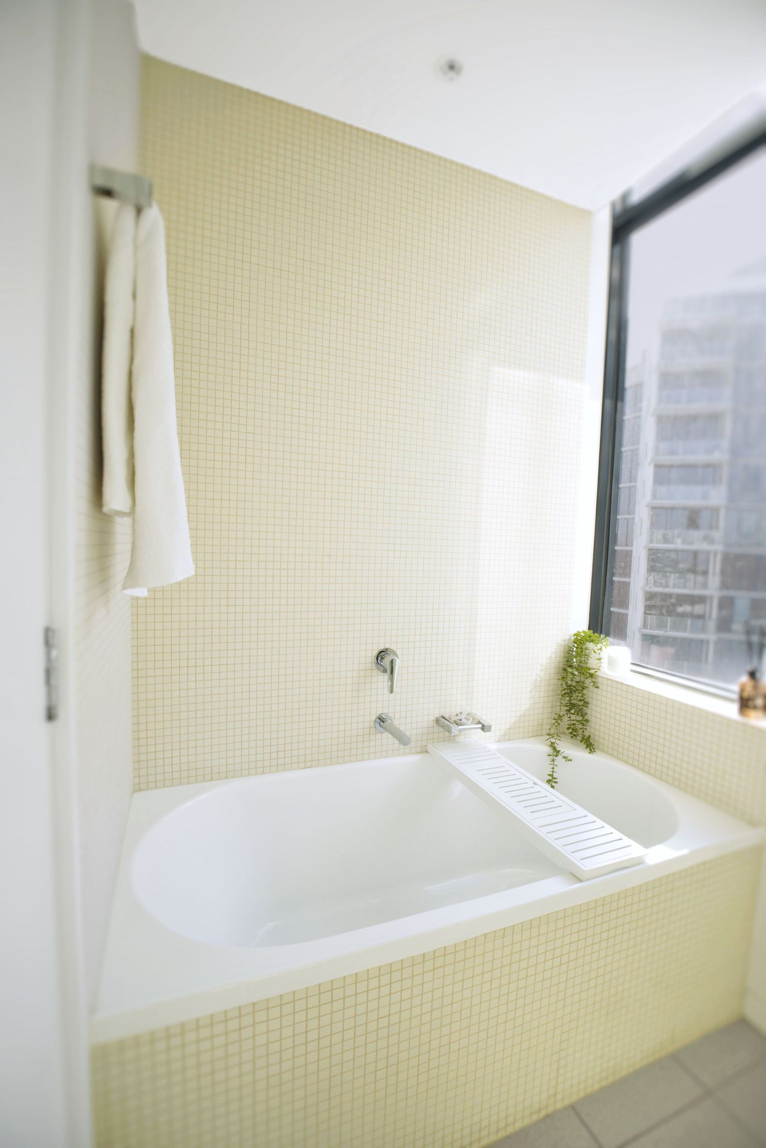 Built-in white bathtub surrounded by cream mosaic tiles, with a bath caddy, hanging towel and corner window.