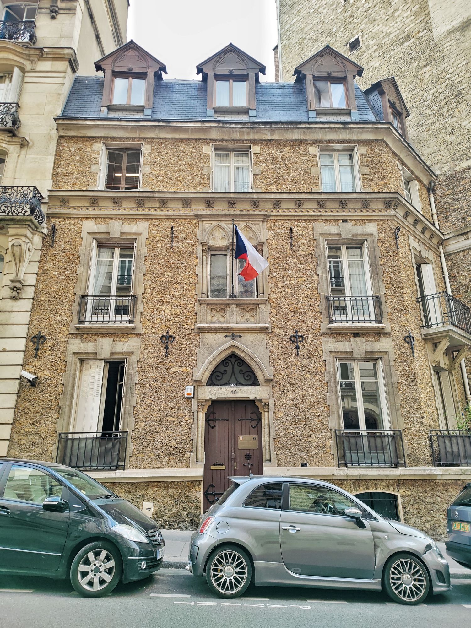 Stone and brick townhouse with a slate mansard roof, dormer windows, a French flag and a gothic arched wooden door on a Paris street.