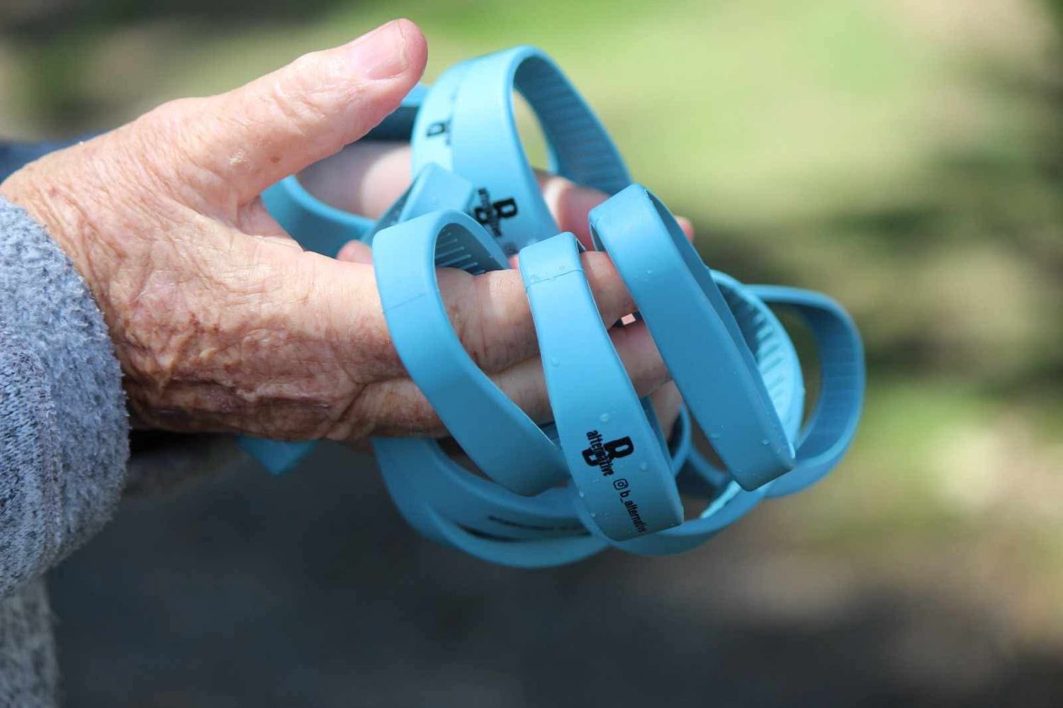 An elderly hand holds several light blue silicone wristbands printed with small black logos against a blurred green background.