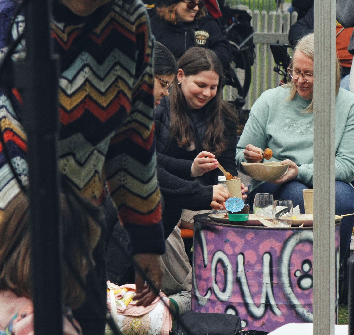 Smiling friends sit around a decorated outdoor table sharing food at a lively community festival.