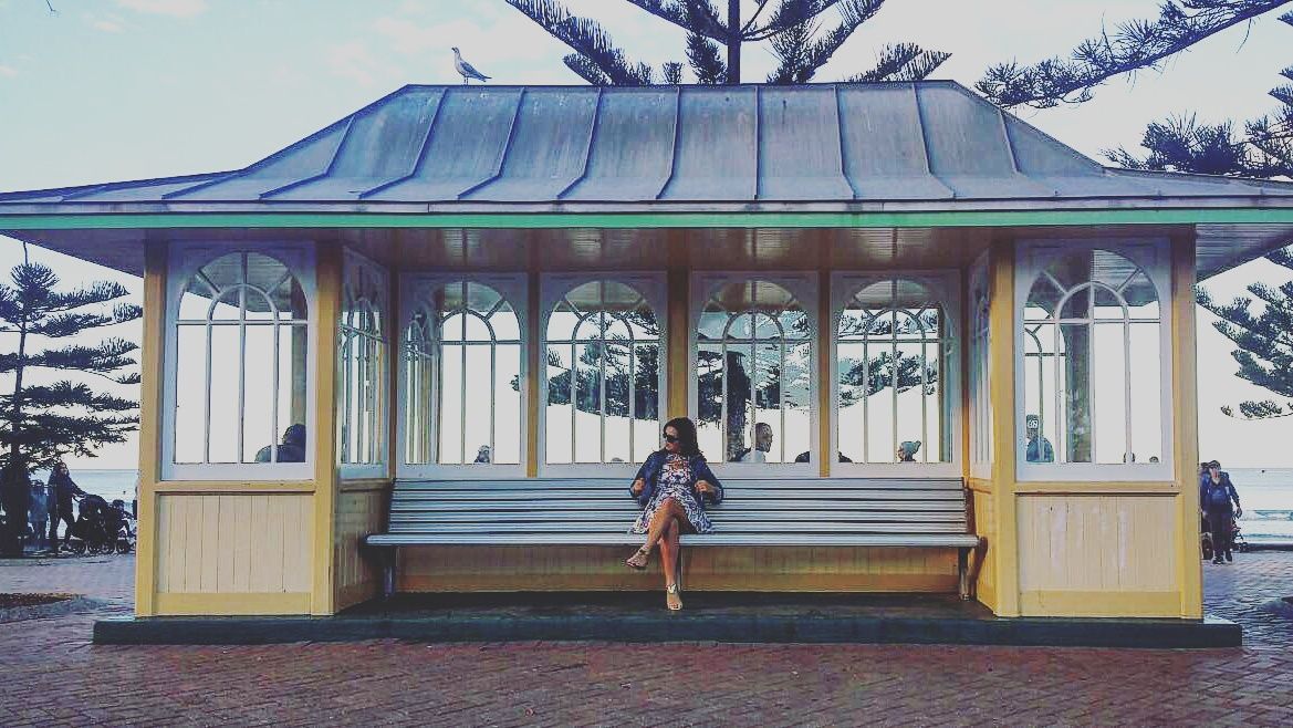A woman sits on a bench inside a historic seaside shelter with a green metal roof, framed by tall Norfolk pine trees.