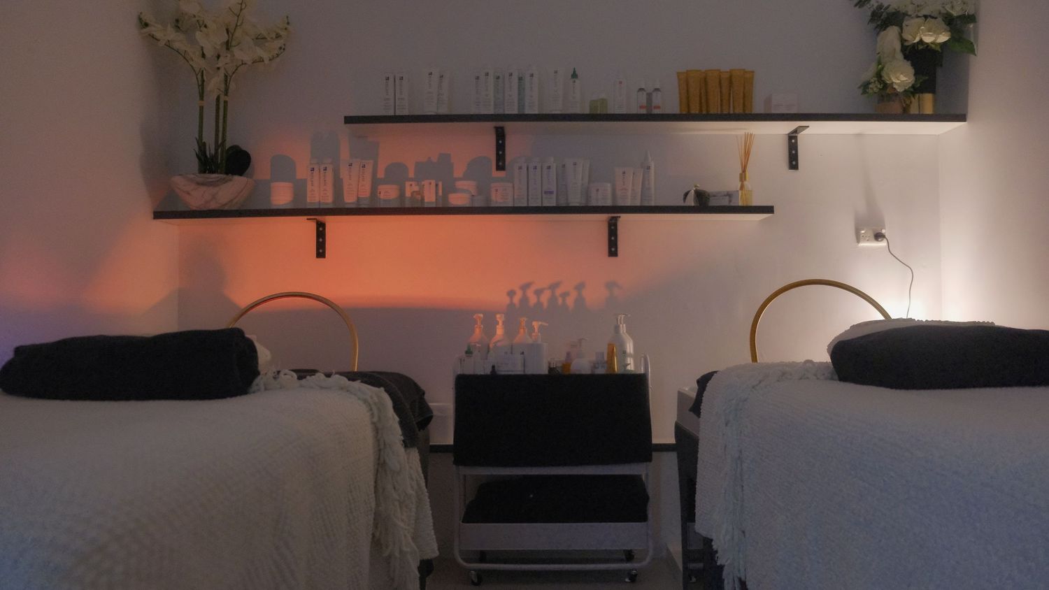 Relaxing spa treatment room with orange ambient lighting, two massage beds, skincare shelves and white flower arrangements.