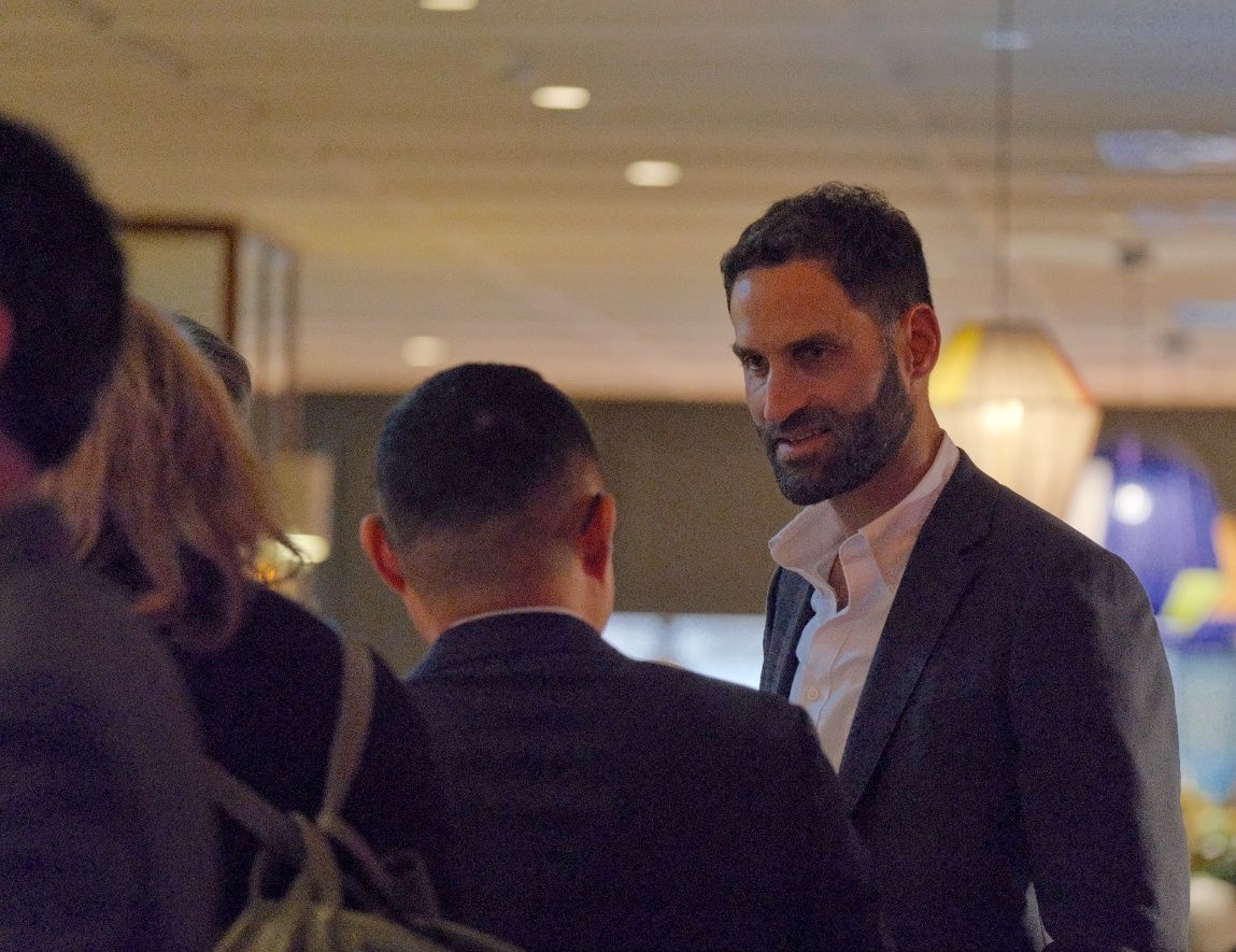 A bearded man in a dark blazer smiles while chatting with guests at an indoor networking event.