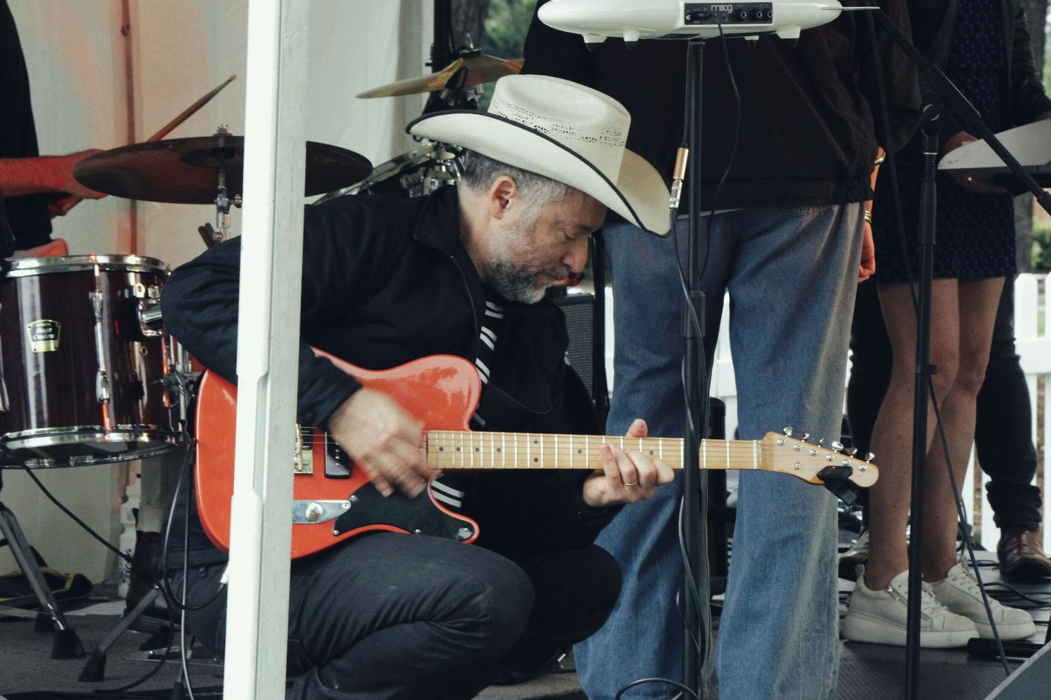 A guitarist in a white cowboy hat crouches on stage playing a red electric guitar beside a drum kit.
