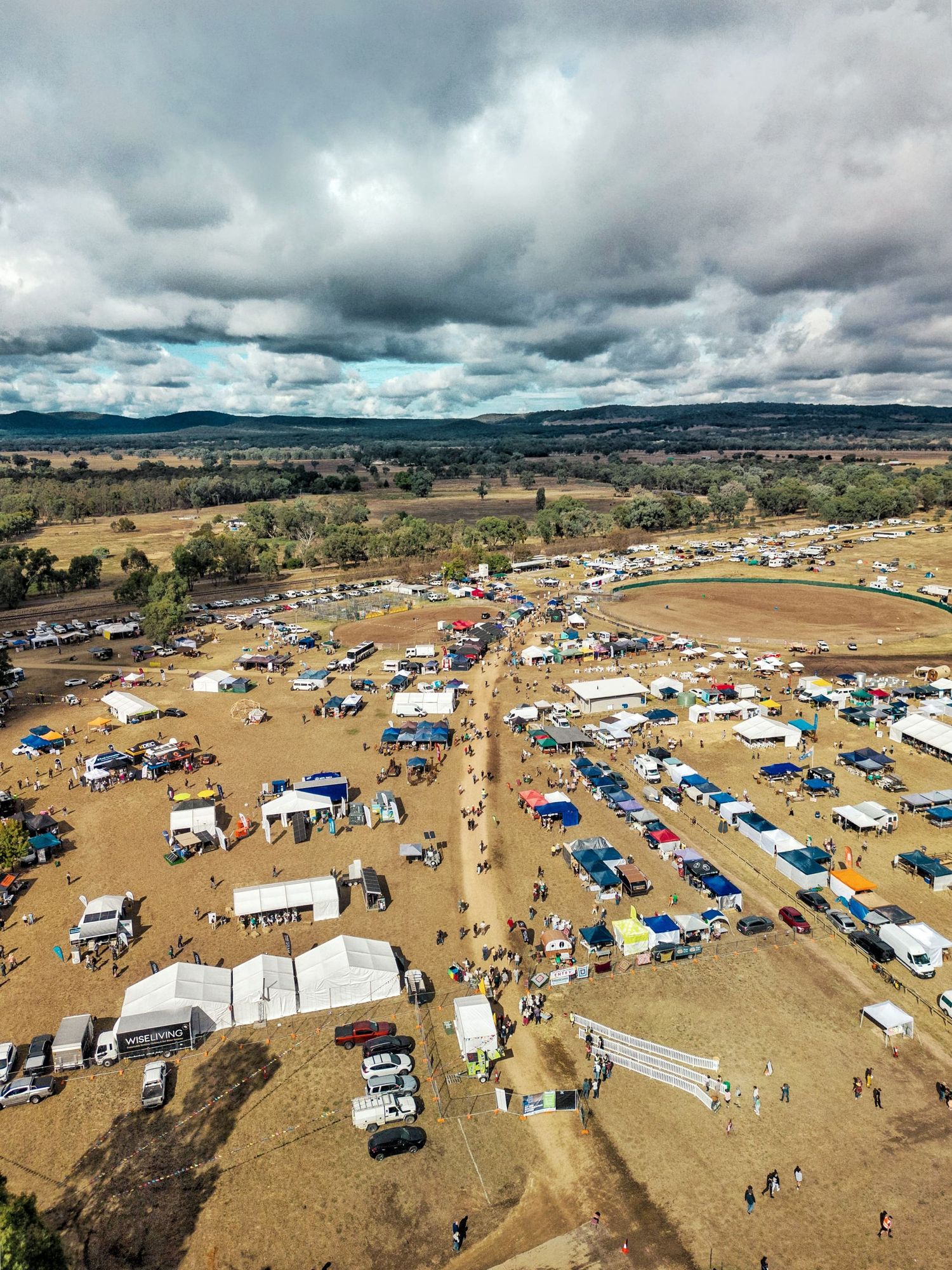 Aerial view of a busy outdoor fair with tents, parked vehicles and crowds on dry ground beneath a cloudy sky and distant hills.