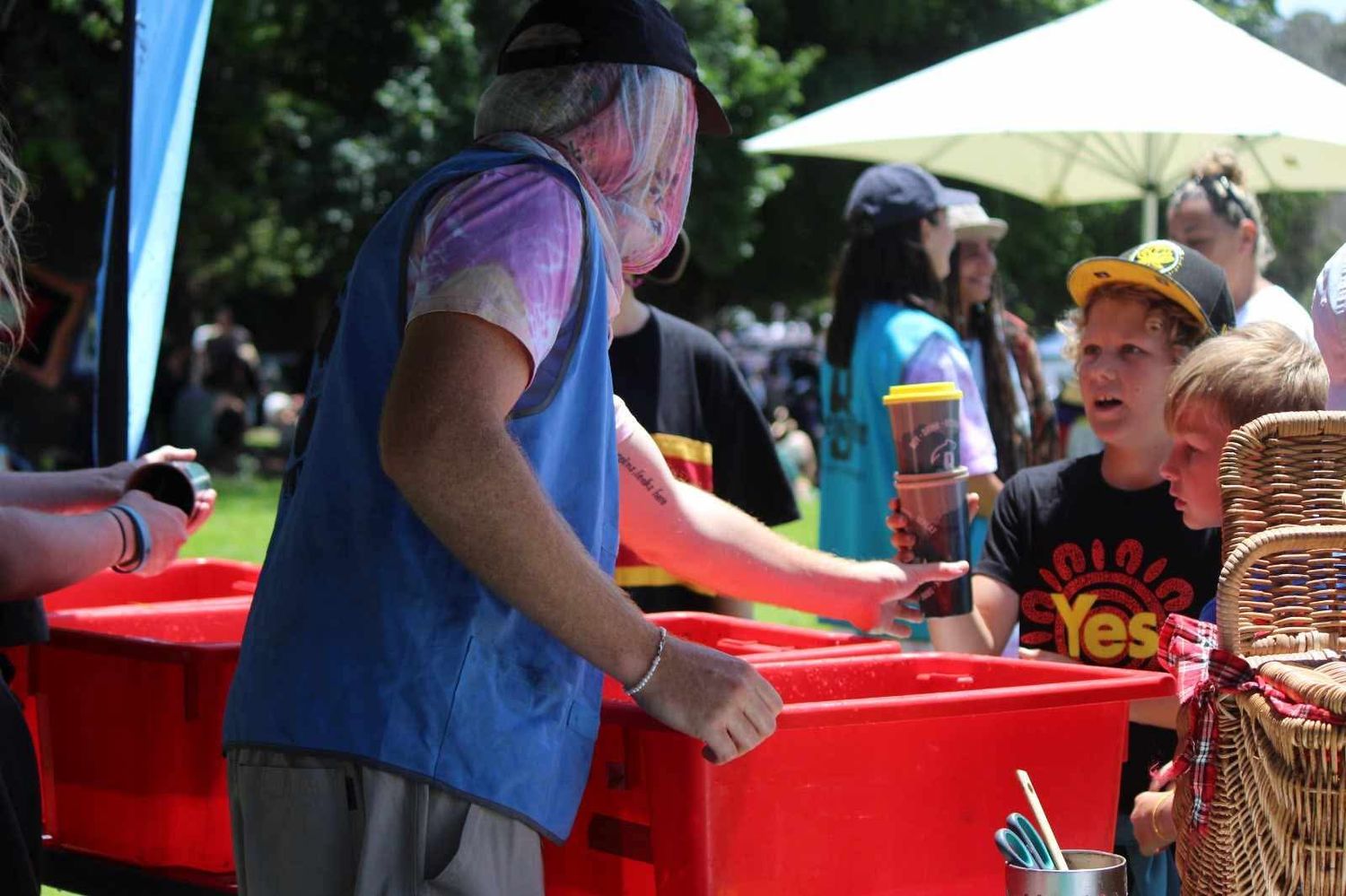 A person in a blue vest serves a boy in a yellow cap at a red table during an outdoor community event with umbrellas and trees.