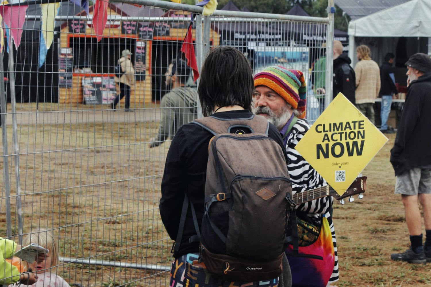 Two festival-goers stand by a fence, one wearing a backpack and the other a striped beanie and holding a yellow Climate Action Now sign.
