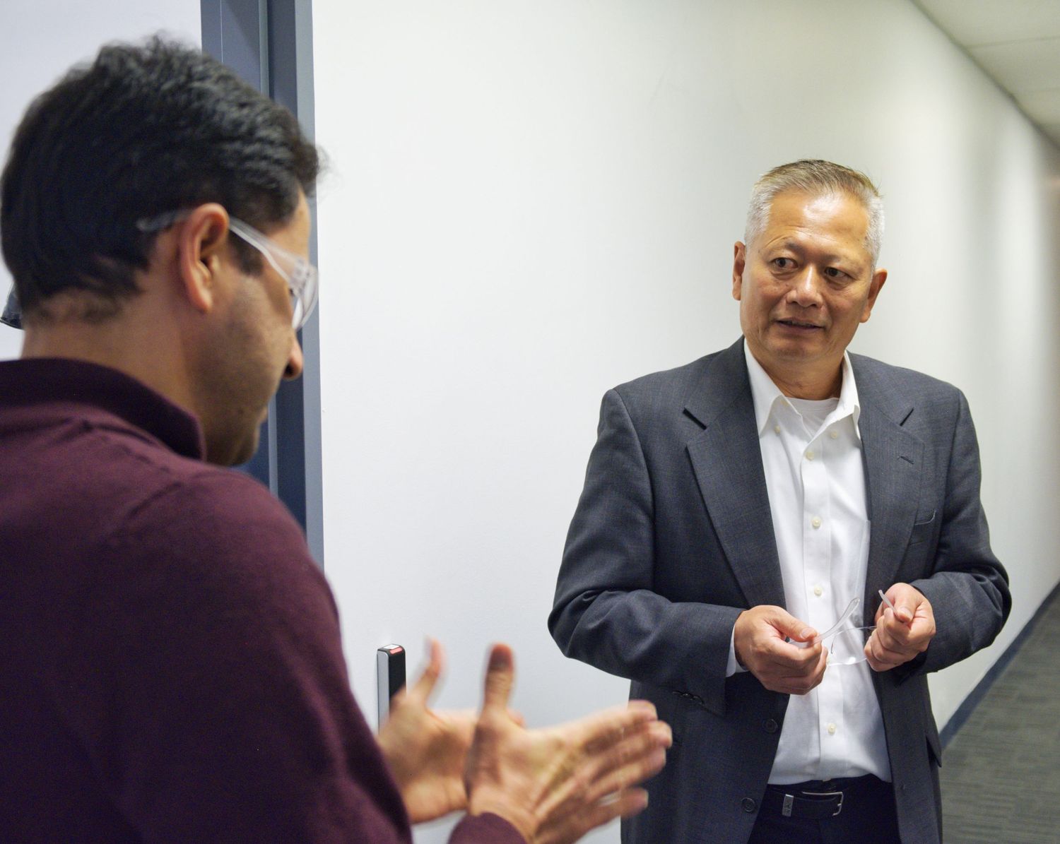 A man in a gray blazer speaks with a colleague in a maroon sweater who gestures in a bright corridor.