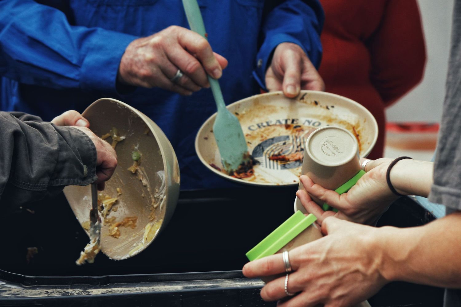 Hands holding used plates, bowls and a green spatula over a tray of leftover food at a communal meal.