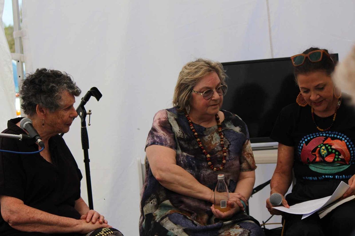 Three women sit on a panel with microphones at an outdoor festival tent, with one holding a bottle and another reading notes.