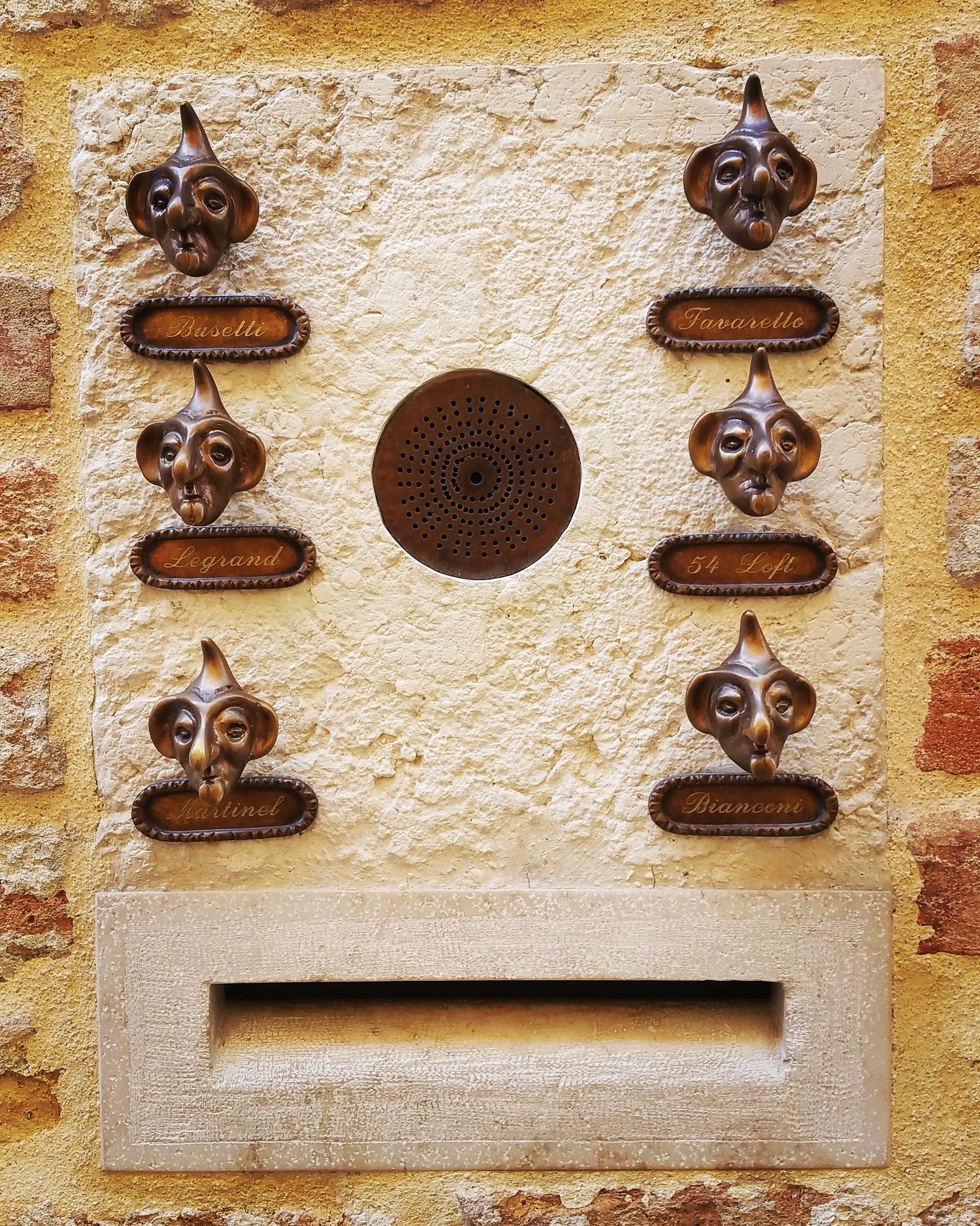 Rustic stone wall intercom panel with six bronze face-shaped bell buttons, name plates and a mail slot.