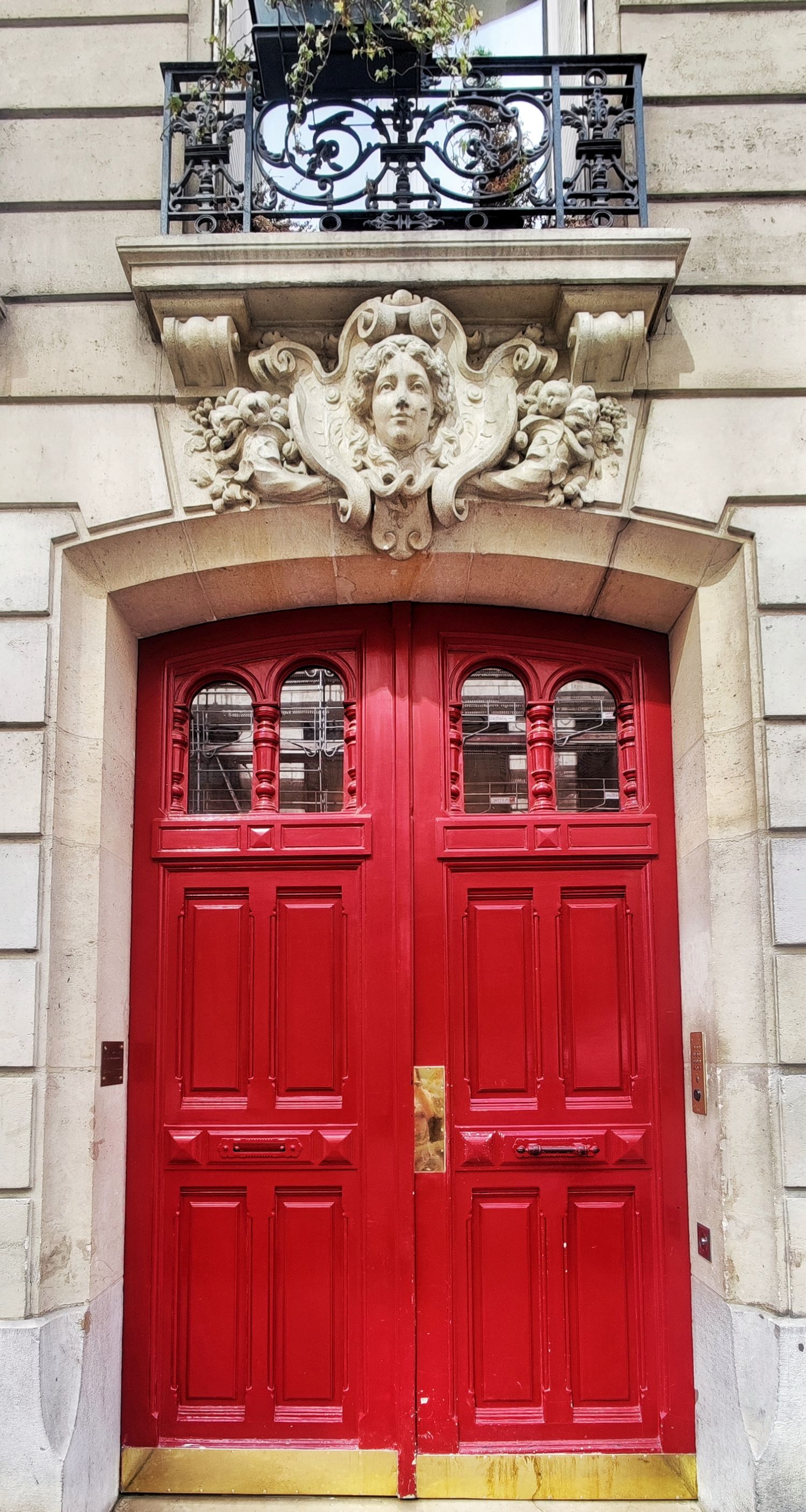 A vivid red double door sits beneath a sculpted stone face relief and a wrought iron balcony on an elegant Parisian building.