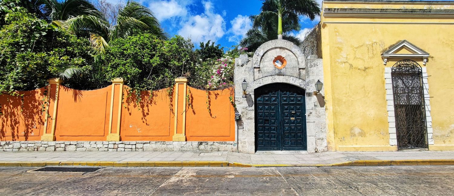 Colonial-style street with a yellow building, ornate stone archway and dark blue double doors beside an orange wall and lush tropical trees.