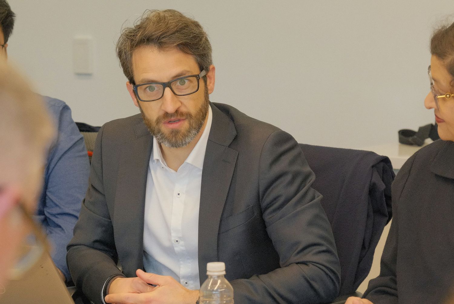 A bearded man in glasses and a dark blazer listens attentively during a meeting, with a water bottle on the table in front of him.