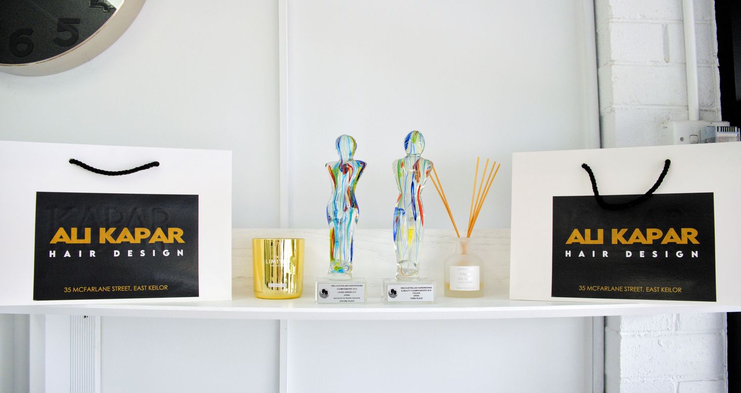 Two Ali Kapar Hair Design shopping bags with glass figurines, a gold candle and a reed diffuser on a white shelf.