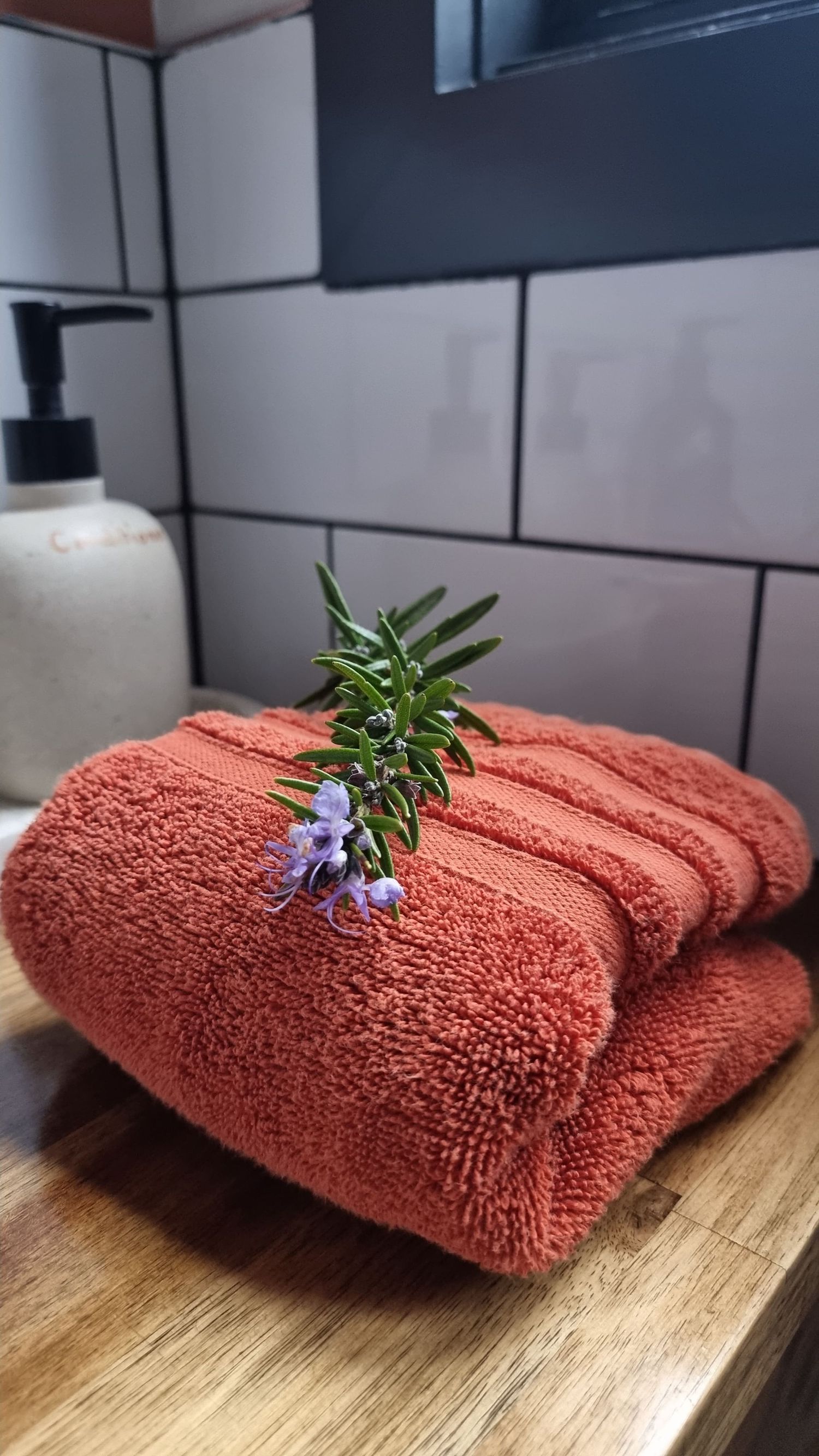 Folded rust-orange bath towel topped with a fresh sprig of purple-flowering rosemary on a wooden bathroom counter beside a soap dispenser.