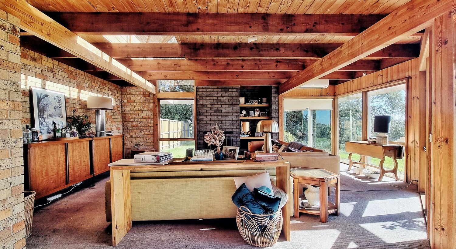 Rustic cabin living space with timber beams, brick walls, wooden cabinetry, a large work table, and big windows with garden views.