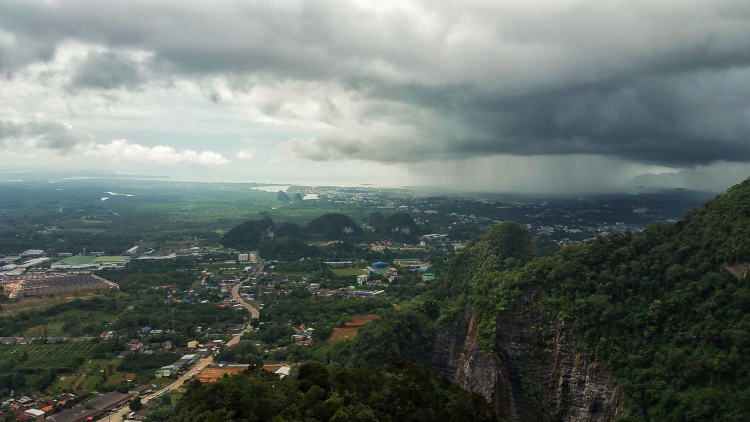 Aerial view of a town and green valley beneath dark storm clouds, with a rocky limestone cliff in the foreground.