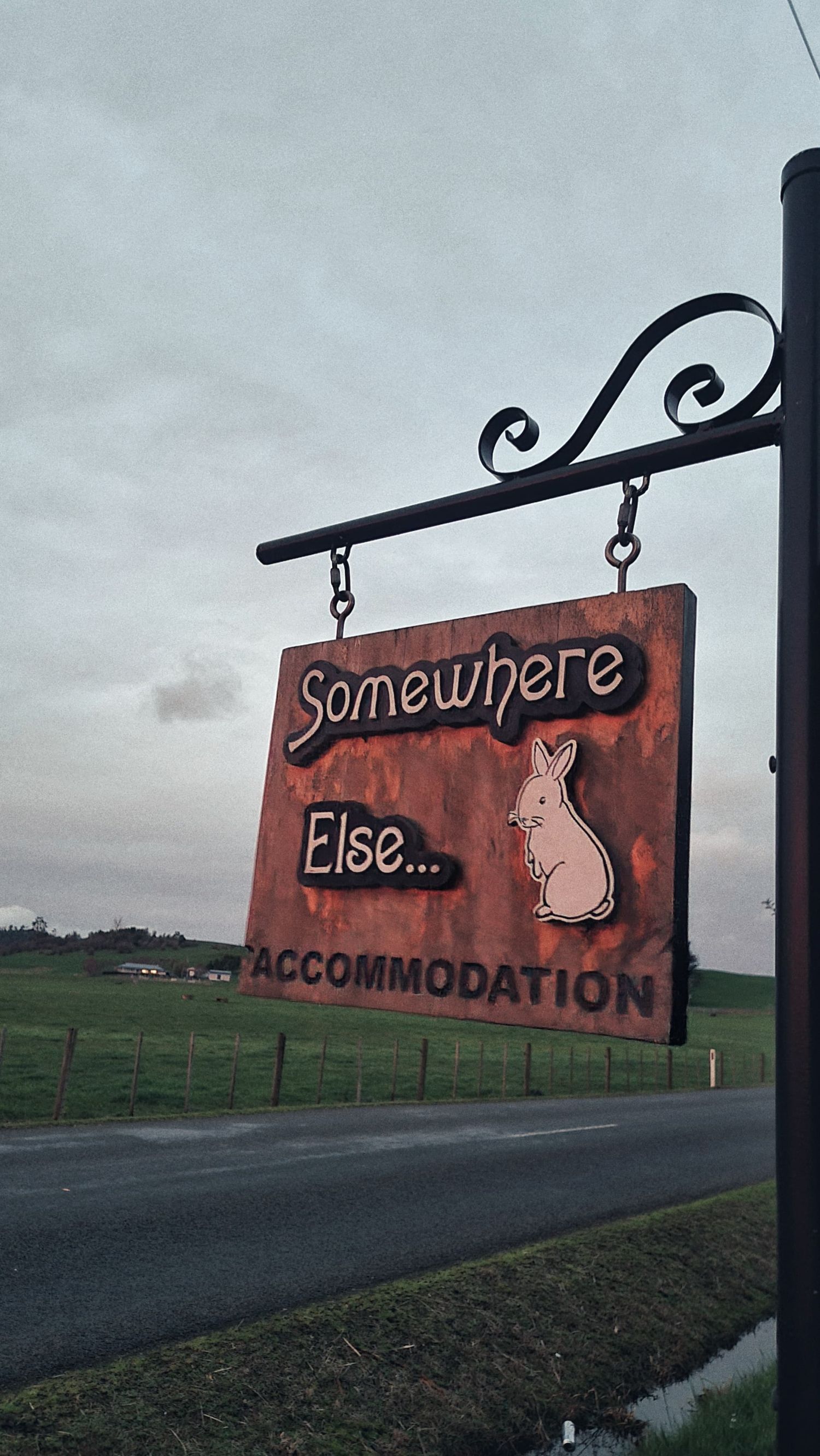 Rustic hanging sign reading Somewhere Else Accommodation with a rabbit carving beside a country road and green rolling hills.
