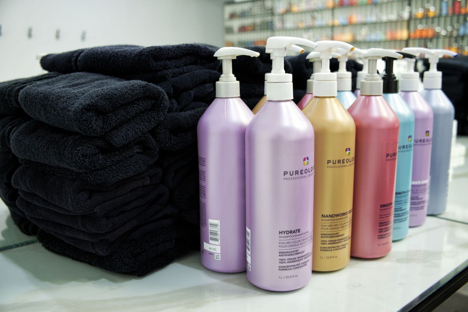 Pureology shampoo and conditioner pump bottles in purple, gold, pink and blue beside a neat stack of black salon towels.