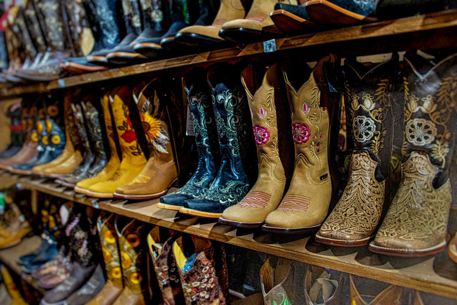 Rows of handcrafted cowboy boots with ornate embroidery and floral designs in tan, yellow, and blue displayed on wooden shelves.