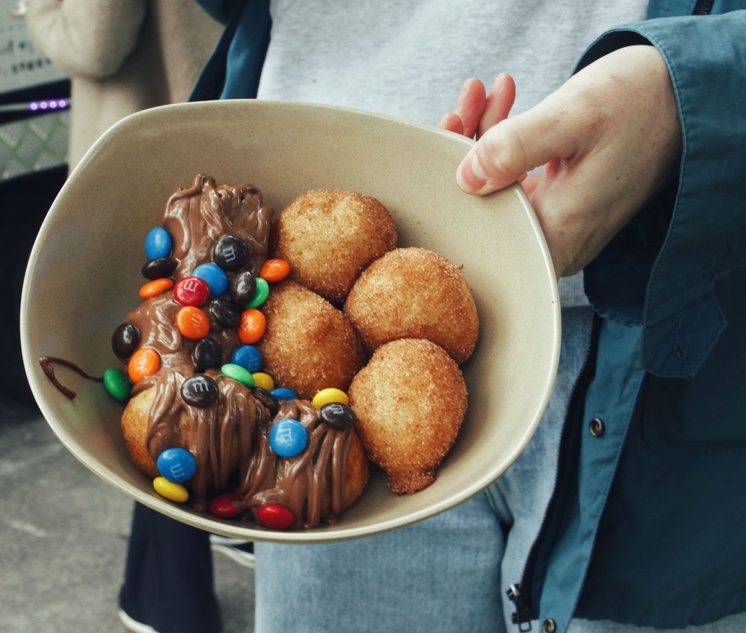 A person holds a beige bowl filled with golden cinnamon doughnut balls and chocolate-sauce treats topped with colourful candy pieces.
