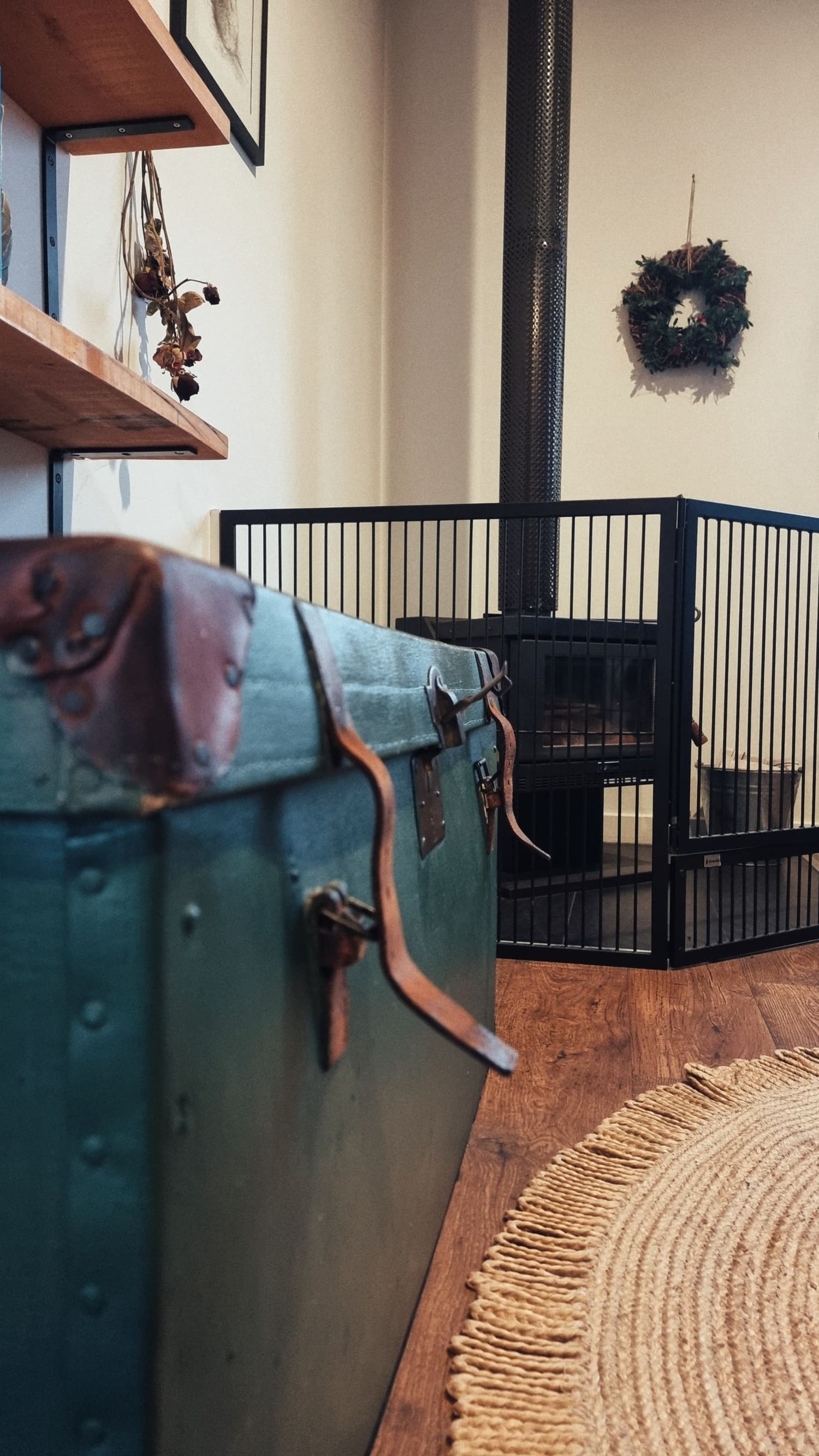 A vintage green steamer trunk sits in a cosy cottage room with timber shelves, a wreath, a black pet gate and a woven round rug.
