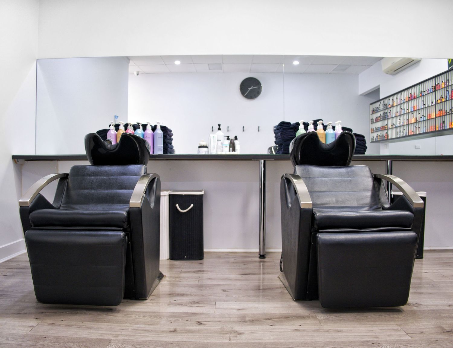 Two black reclining shampoo chairs in a modern hair salon with a mirror, counter and hair product bottles.