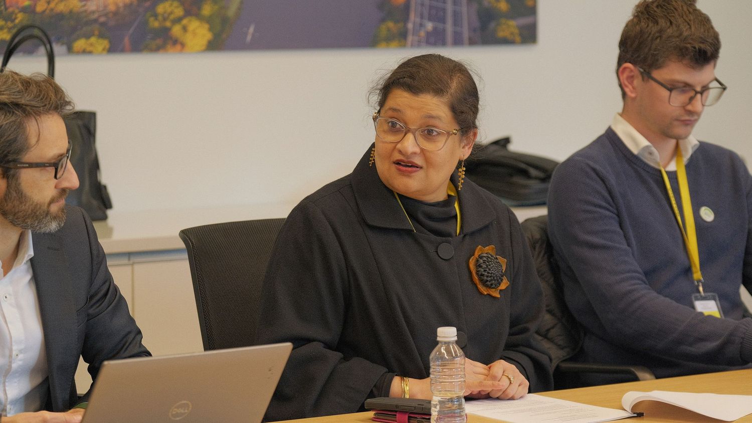 A woman in a dark jacket speaks at a conference table with two men beside her, one using a laptop.