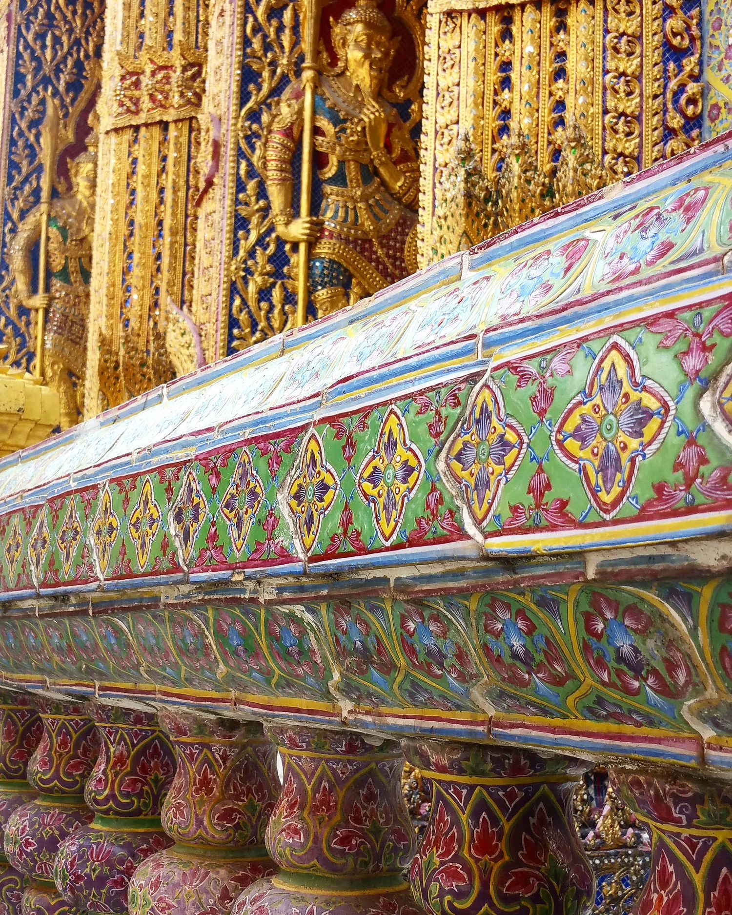 Ornate Thai temple detail with colorful glazed ceramic tile patterns, gilded carvings and a golden guardian figure.