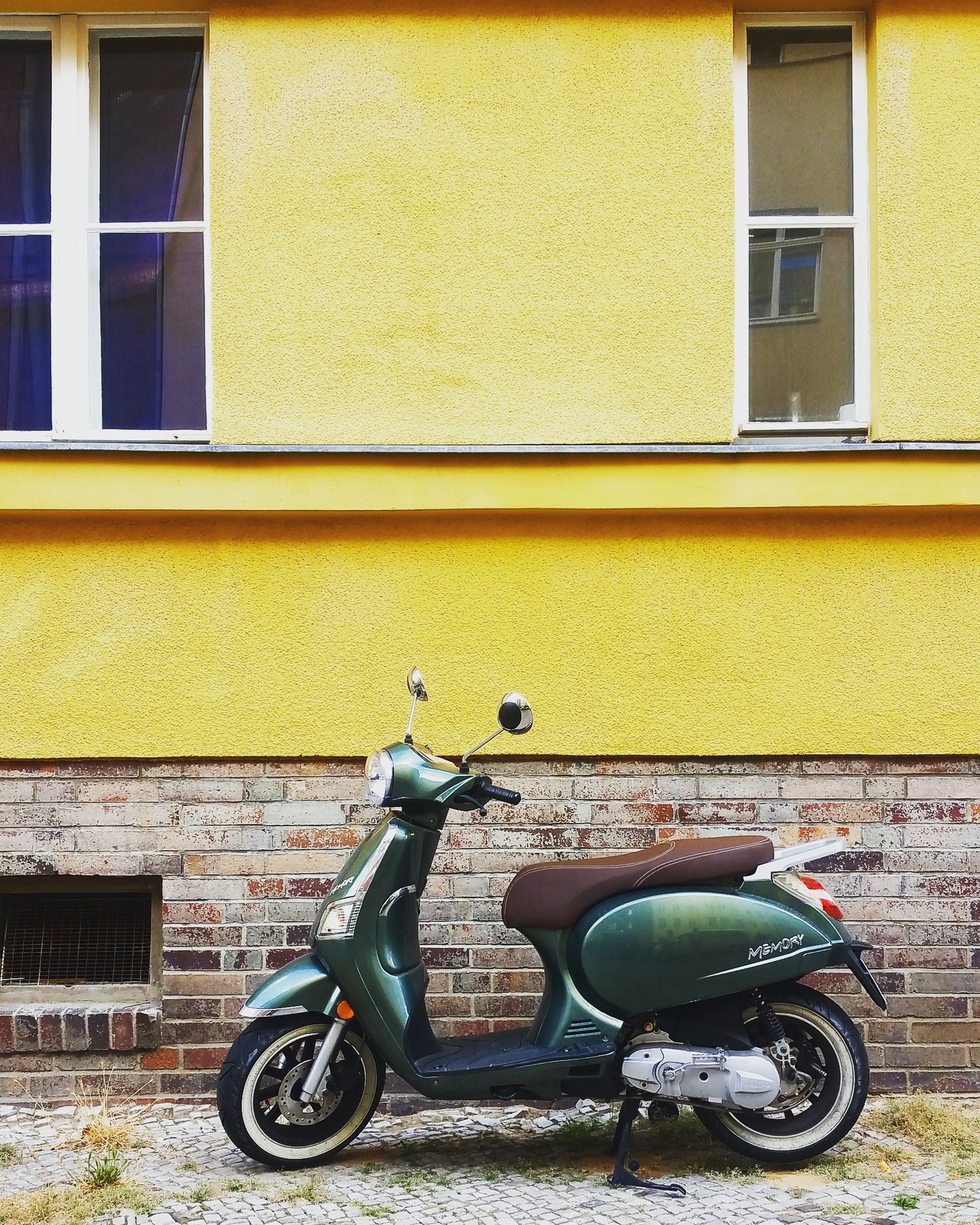 Green vintage scooter parked in front of a yellow stucco wall with a brick base and two windows.