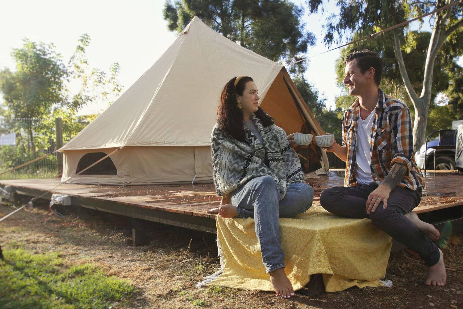 Couple sitting and laughing on a yellow blanket in front of a glamping bell tent on a timber platform in bushland.