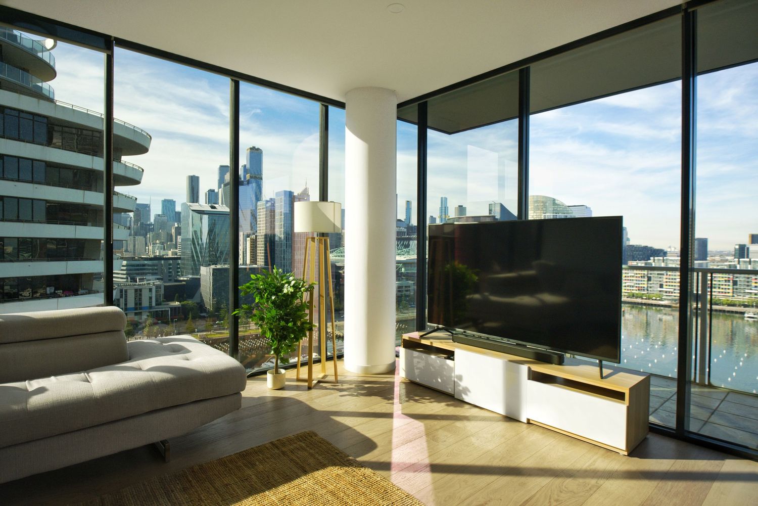 Bright apartment living room with floor-to-ceiling windows, a sofa, flat-screen TV and sweeping city skyline and water views.