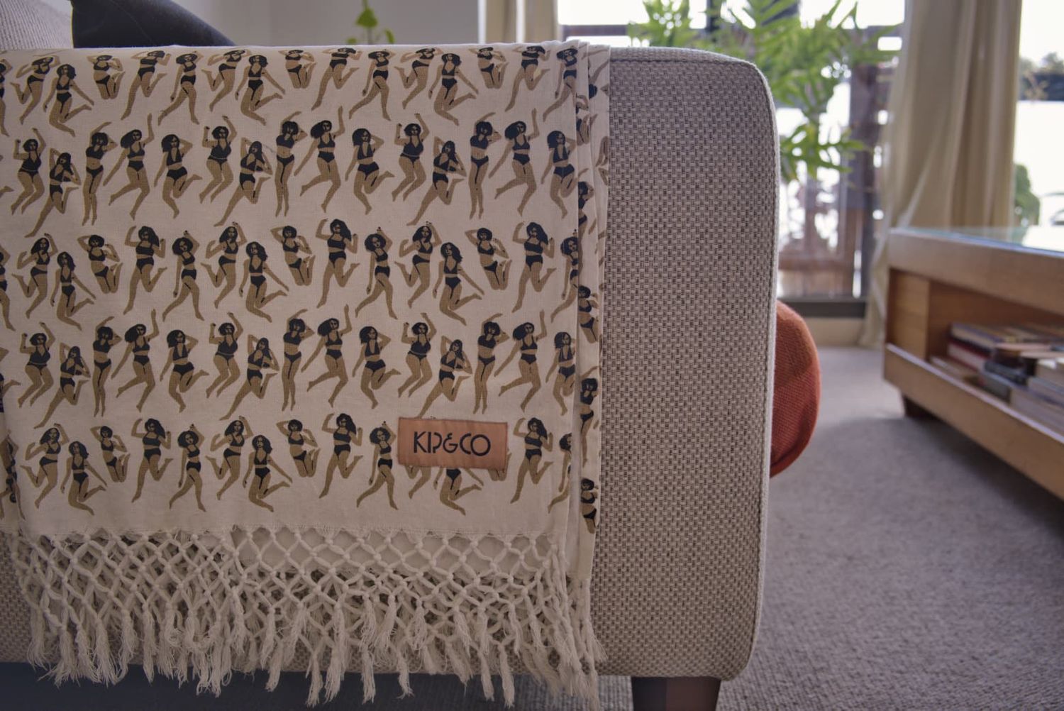 Close-up of a cream fringed throw blanket with a printed figure pattern draped over the arm of a sofa.