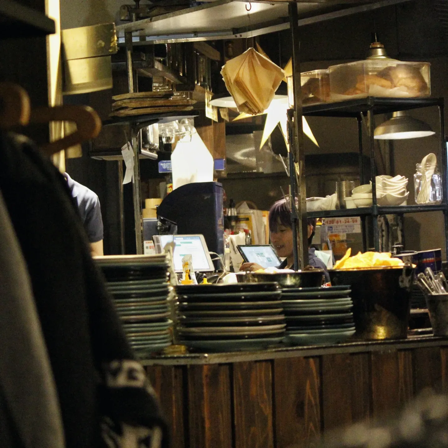 Cozy Japanese izakaya kitchen counter with stacked plates and cookware as a staff member works in the background.