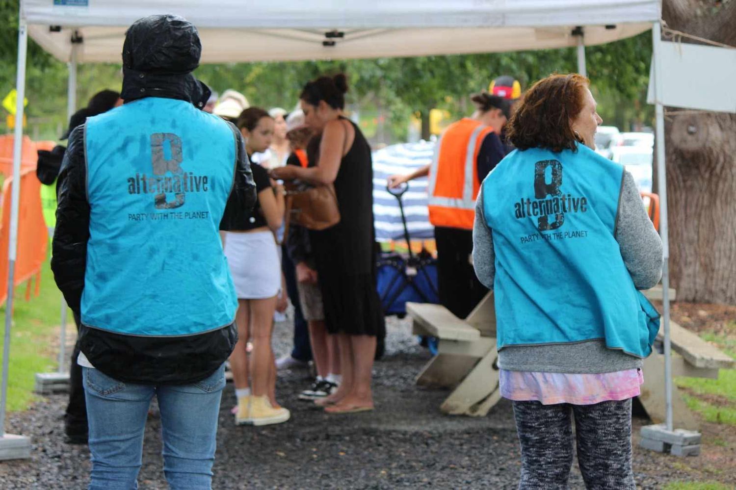 Two volunteers in teal vests stand beneath a white marquee at a community event while people gather around a table in the background.