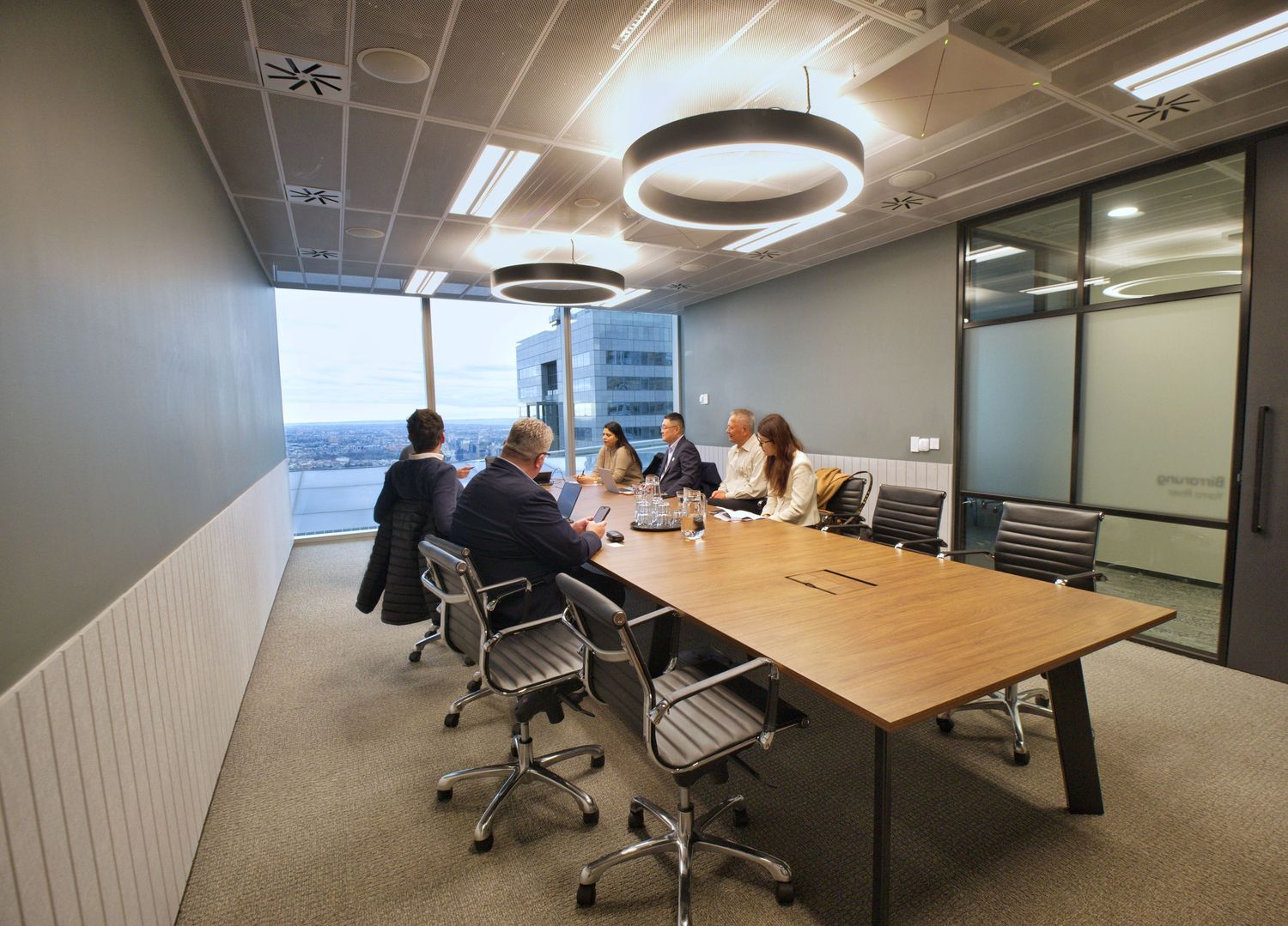 A small group of colleagues meets around a wooden table in a bright modern conference room with large windows and city views.