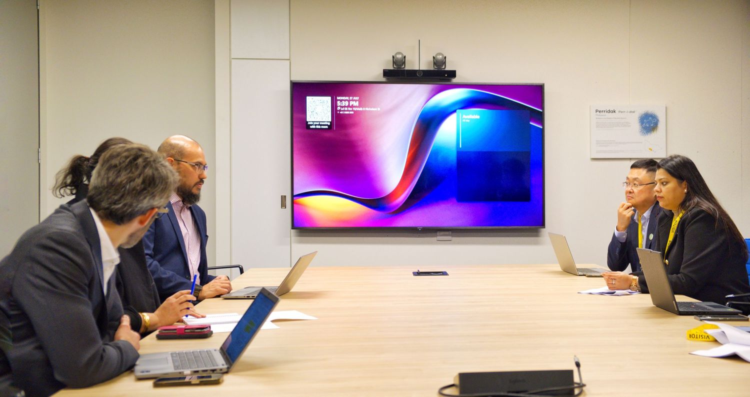 Colleagues with laptops sit around a light wood conference table facing a large wall screen showing a colorful abstract display.