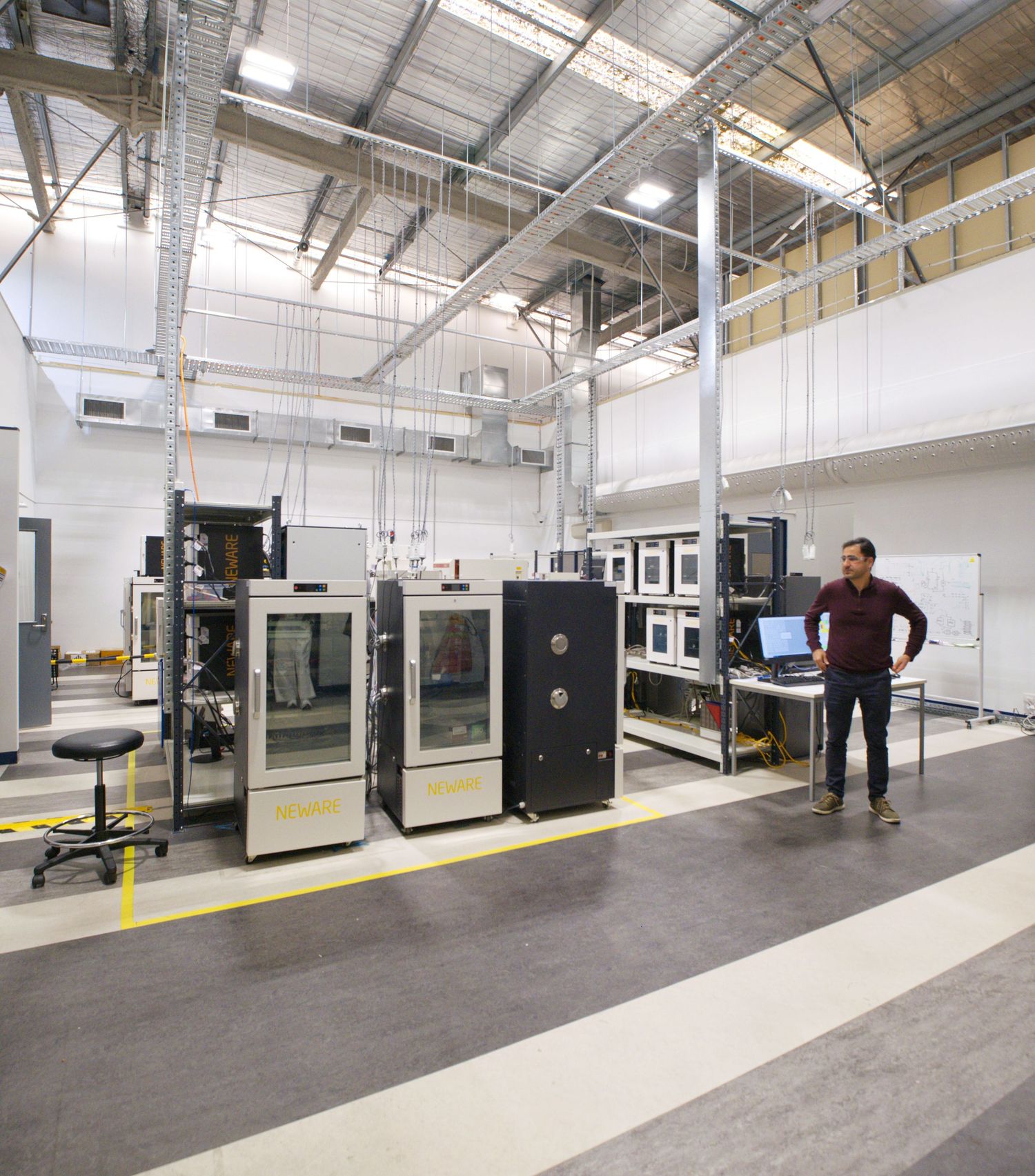 A man stands in a spacious battery testing laboratory with environmental chambers, workstations and exposed ceiling beams.