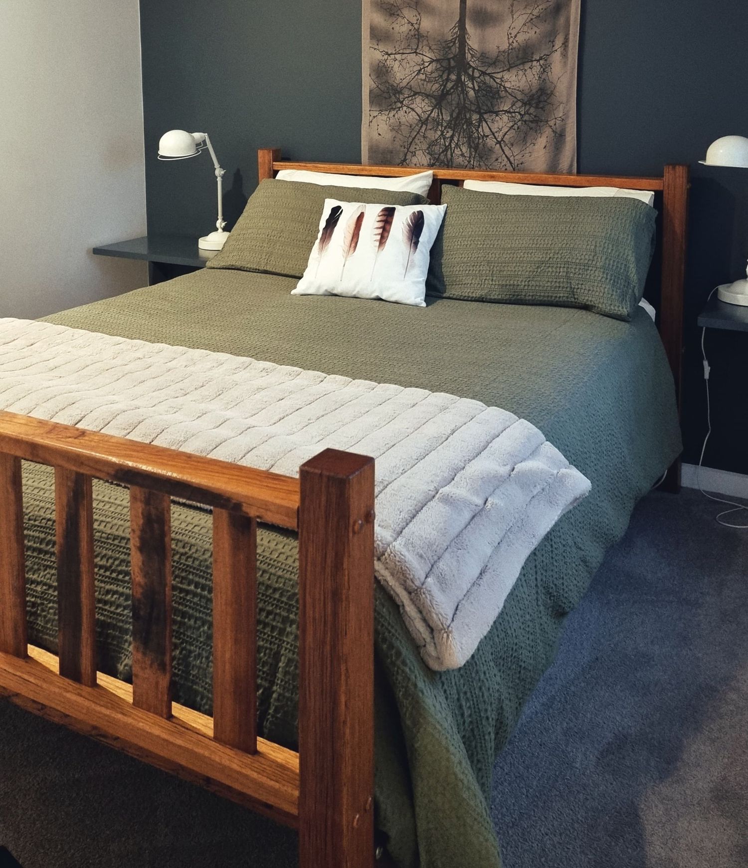 A queen bed with olive green linen, a chunky knit throw and a rustic timber frame stands against a charcoal feature wall.
