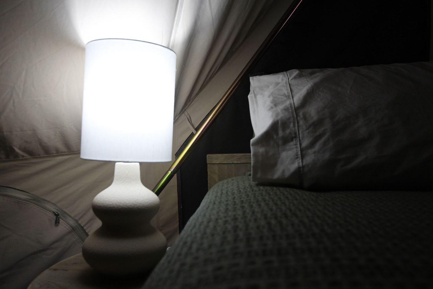 A softly glowing white table lamp sits beside a bed with grey bedding inside a canvas glamping tent at night.