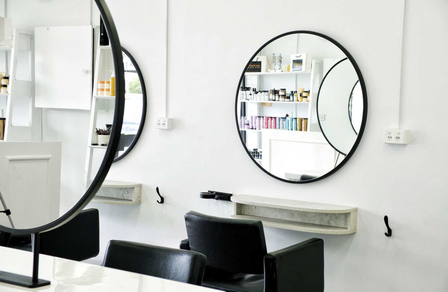 Close view of round black-framed salon mirrors on white walls above styling stations with black chairs and shelves of hair products.