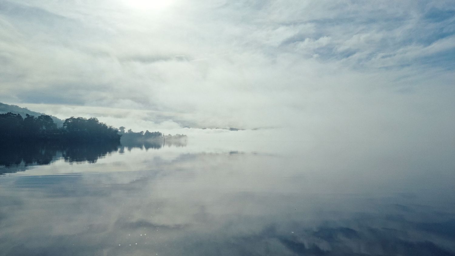 Soft morning mist hangs over a calm lake, with clouds reflected in the still water and a dark tree-lined shoreline in the distance.
