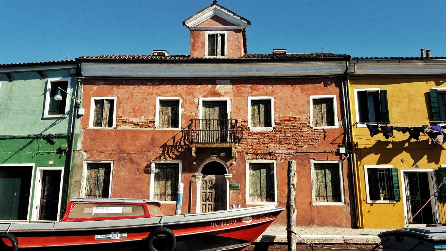 Weathered red and yellow Burano houses with green shutters and a red boat moored along the canal.