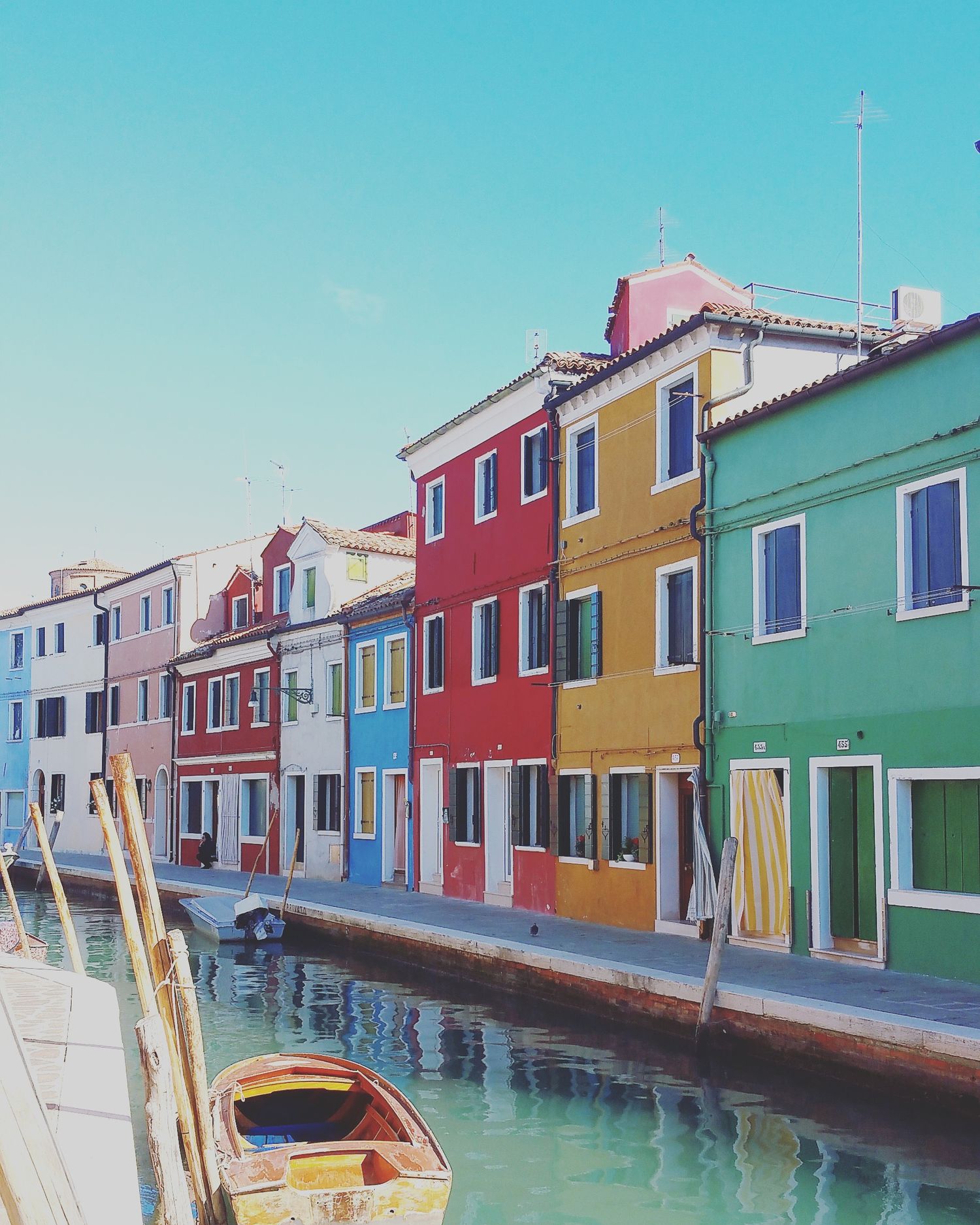 Colorful red, yellow, green and blue houses line a calm canal in Burano with a small wooden boat in front.