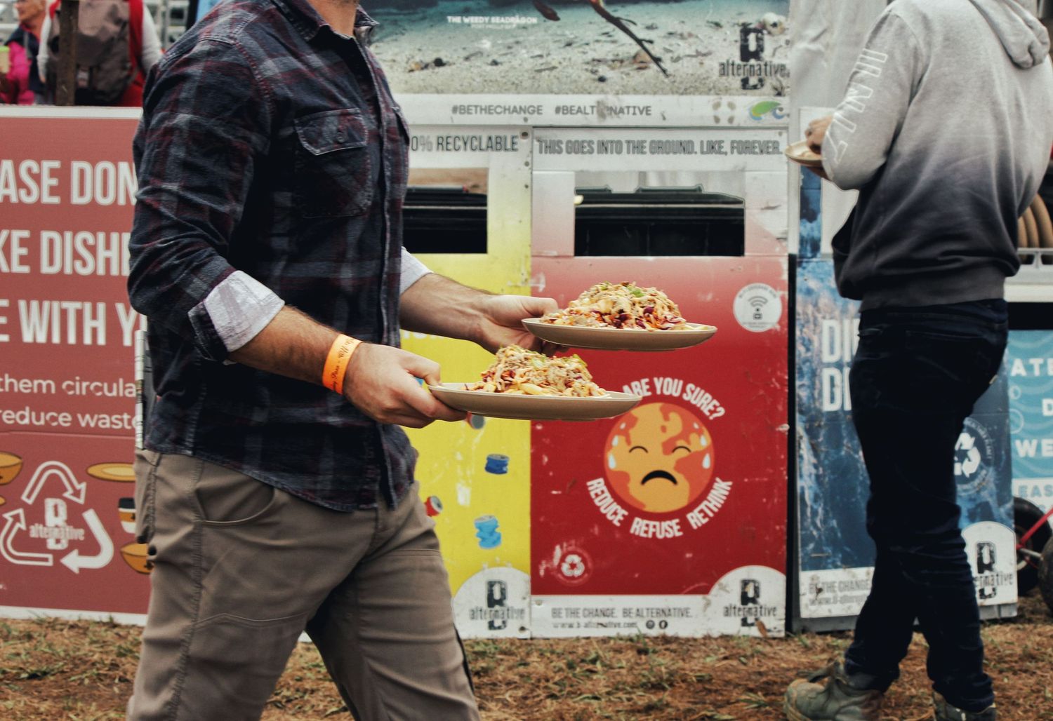 Festival attendee in a plaid shirt carries two plates of food past colorful signs promoting waste-free dining.
