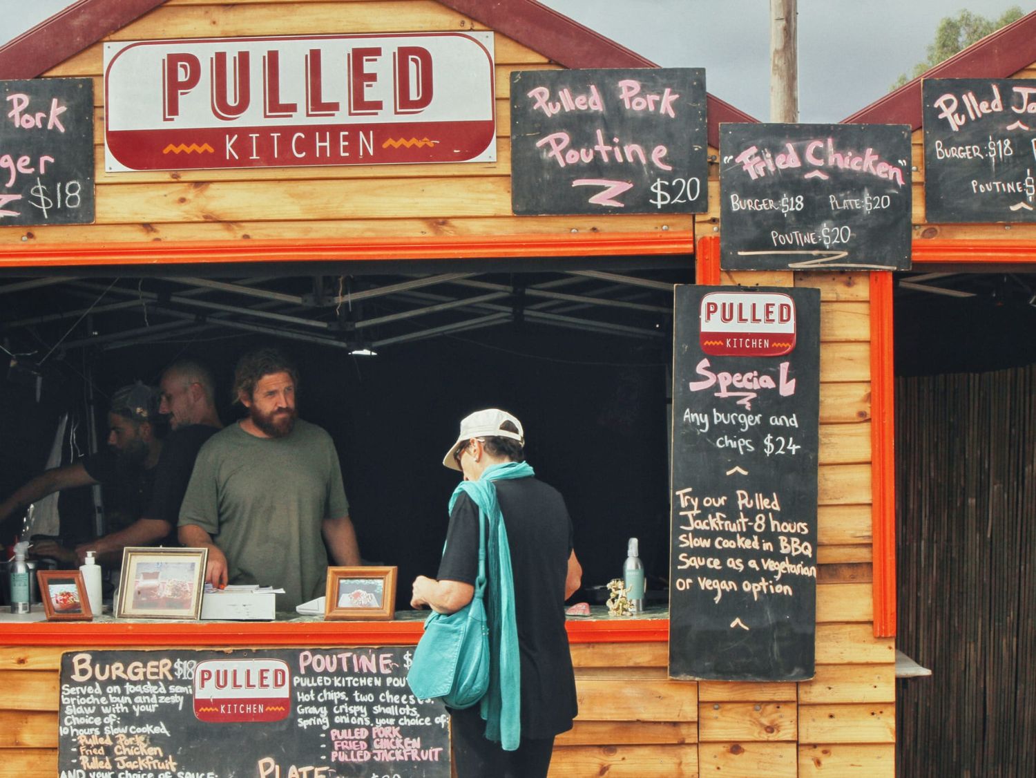 Visitors stand at the Pulled Kitchen food stall, where chalkboard menus list pulled pork, poutine and fried chicken at a festival.