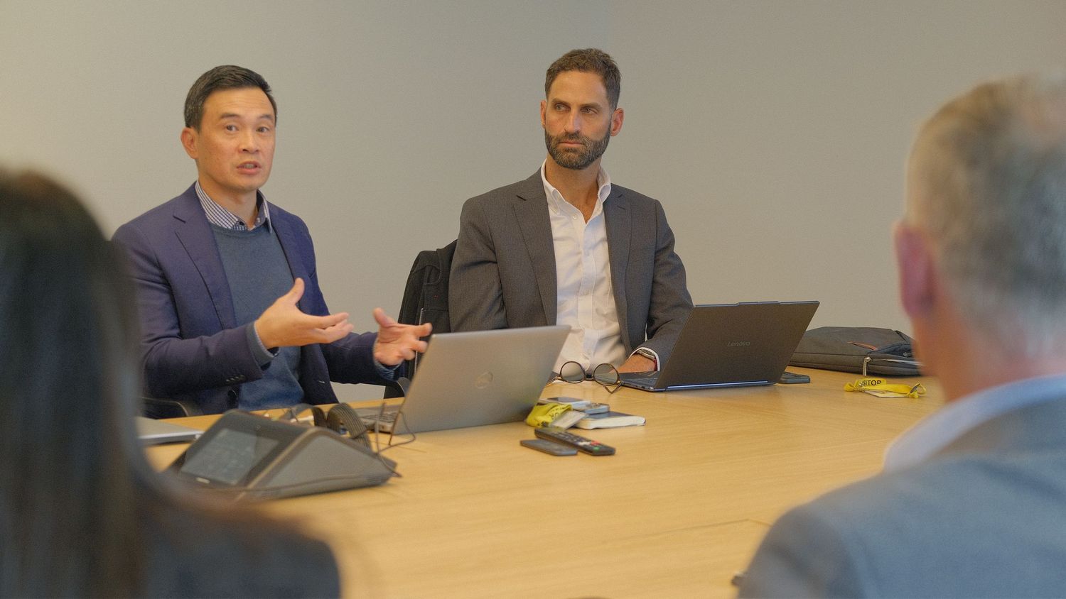Two businessmen sit side by side at a conference table with laptops, one gesturing as he speaks during a team meeting.